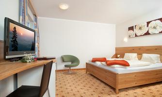 A modern bedroom with a large bed, a desk, and a television. The walls are light, and the decoration consists of painted artwork and a floral carpet.