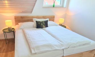 A cozy bedroom with a double bed and light wood paneling. Next to the bed, there are two lamps and a window lets in plenty of daylight.