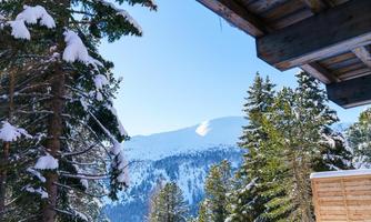 A winter landscape with snow-covered mountains and green fir trees. The sky is clear and blue, and you can see a wooden balcony in the foreground.