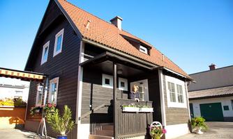 A beautiful wooden house with a red roof and a porch. In front of the house, there are flowers and plants, and the sun is shining.