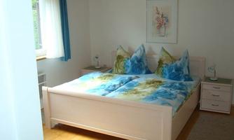 A cozy bedroom with a double bed and colorful cushions. The room features a light wooden floor and large windows that let in plenty of daylight.