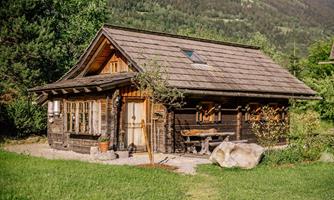 A charming wooden house amidst green meadows and trees. Gentle hills and a tranquil landscape are visible in the background.