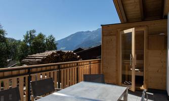 A balcony with a table and chairs, surrounded by trees and mountains. The sky is clear and blue, creating a relaxing atmosphere.