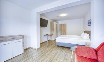 A modern hotel room with a double bed, a television, and a red sofa. The spaces are bright and friendly designed, with wooden flooring and minimal decoration.