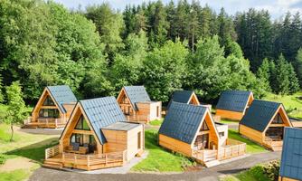 Modern wooden buildings in a green environment. The small cabins have sloped roofs and are surrounded by trees.