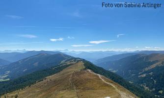 An impressive mountain landscape with gentle hills and a wide view of the Alps. The sky is clear and blue, ideal for hiking.