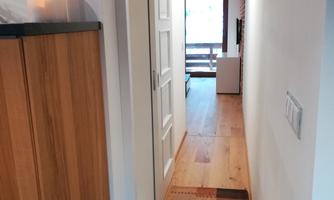 A bright corridor with wooden flooring and a colorful rug mat. At the end of the corridor is an open door that leads to a living area.