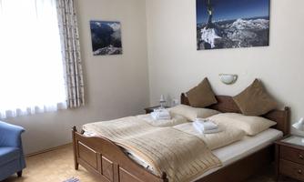 A cozy bedroom with a double bed, covered with white bedding and towels. In the background, an armchair and a window with daylight are visible.