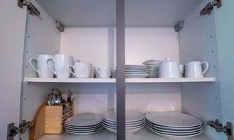 A kitchen cabinet with white plates, cups, and a coffee pot. On the lower shelf, there are more plates and cutlery.