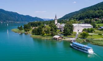 A picturesque lake with clear water and green shores. In the background, mountains and a church can be seen.