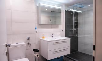 A modern bathroom with a shower and a simple sink. The walls are tiled in a light color, and there is a large mirror above the sink.