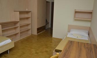 A simple room with two beds, a desk, and shelves. The floor is wooden and the walls are painted light.