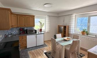 A modern kitchen with wooden cabinets and a bright dining table. Large windows let in plenty of light.