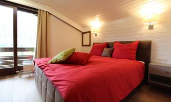 A cozy bedroom with a large red bed and colorful pillows. Large windows provide plenty of daylight and an inviting atmosphere.