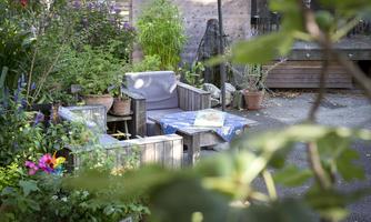 A cozy garden with wooden furniture and colorful plants. In the background, more plants and a statue can be seen.