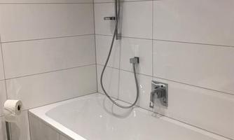 A modern bathroom with a white bathtub and a handheld shower. The walls are tiled, creating a sophisticated, clean ambiance.
