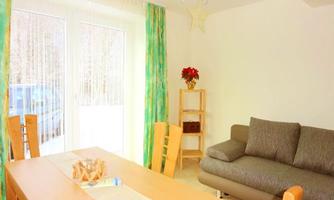 A bright room with a wooden table and chairs. A cushion lies on the sofa, and large windows let in a lot of light.