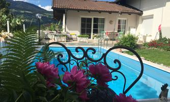 A beautiful pool surrounded by green plants and colorful flowers. In the background, a bright house and the mountains can be seen.