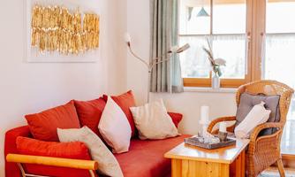 A cozy living room with a red sofa and a wicker chair. A decorative artwork hangs on the wall, while a table with a small decoration stands in the middle.