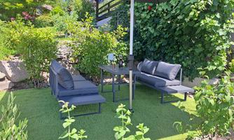 A cozy seating area in the garden with modern sofas and a table.  
Surrounded by green plants and a sunshade.