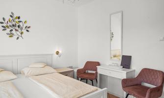 A bright bedroom with two beds, a mirror, and a small table. The accent chairs are in a warm brown tone and the walls are simply white.