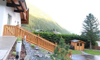 A house with a wooden porch and a beautiful view of the mountains. In the foreground, there is a well-maintained garden with stones and plants.