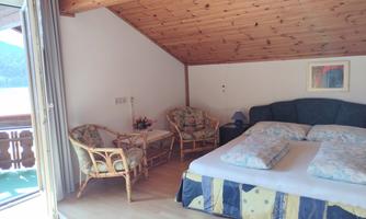 A cozy bedroom with a double bed and two wicker chairs. The wooden roof and the balcony create a warm, inviting atmosphere.