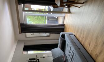 A modern living room with a gray sofa and a dining table. Large windows provide a view of a balcony and plenty of natural light.