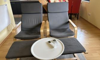 Two modern armchairs with a wooden frame are positioned across from a small table. In the background, a kitchenette and red chairs are visible.