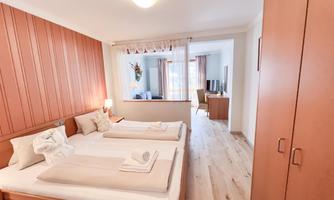 A cozy bedroom with two beds and warm wooden details. In the background, there is a living area with a TV and windows with curtains.