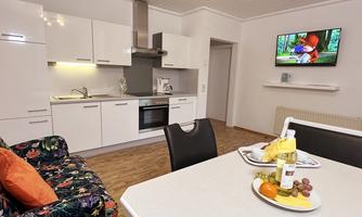 A modern kitchen with bright cabinets and a dining area. A TV is showing a talk show, while fresh fruit is on the table.