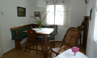 A cozy dining area with a wooden table, chairs, and a bench. A vase with fresh flowers sits on the table, and the room has large windows that let in plenty of light.