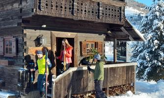 A cozy wooden house in the snow, surrounded by a winter landscape. Three people are preparing for skiing.