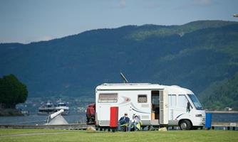 A motorhome is parked on a meadow by the water. In the background, mountains and a boat can be seen.
