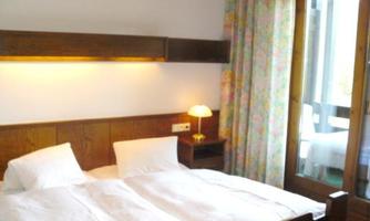 A simple bedroom with two single beds and soft lighting. Windows with curtains let in daylight.