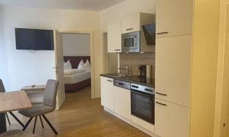 A modern kitchen with appliances and a dining table. In the background, a bedroom with a bed is visible.