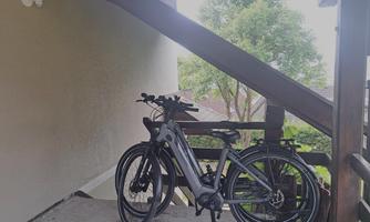 Two e-bikes are standing on a covered terrace. In the background, trees and a quiet residential area can be seen.