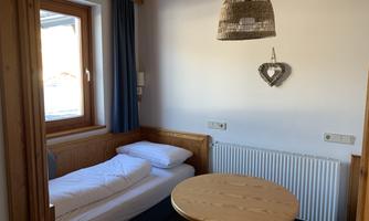 A cozy room with a bed, a round table, and a large window. The wooden style and warm colors create an inviting atmosphere.