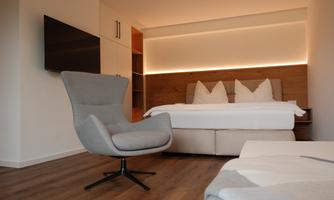 A modern bedroom with a cozy bed and a comfortable chair. The warm wood and light accents create an inviting atmosphere.
