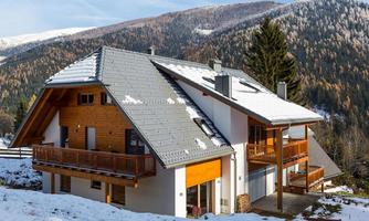 A modern house in the snow, surrounded by mountains. The facade is partly made of wood, and there is a large balcony.