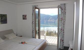 A cozy bedroom with two beds and colorful curtains. The large window front offers a beautiful view of a lake and the surrounding mountains.
