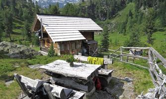 A cozy cabin in the middle of the mountains, surrounded by green meadows and trees. In the foreground, wooden tables and chairs are set up on the grounds.