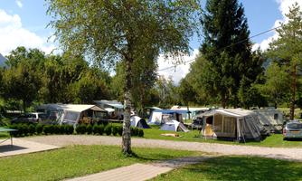 A campsite with several tents and RVs in a green setting. In the background, trees and mountains are visible, the sky is clear and sunny.