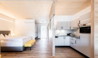 A modern room with a large bed and a kitchenette. The room has bright walls and a friendly wooden floor.