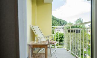 A cozy balcony with a lounge chair and a small table. The view shows green trees and a clear sky.