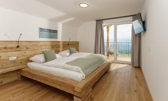 A modern bedroom with a wooden bed and two pillows. The room has large windows overlooking the landscape and a television on the wall.