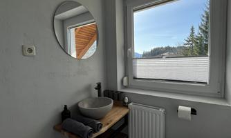 A modern bathroom with a round mirror and a stone sink. Through the window, there is a beautiful view of trees and a clear sky.
