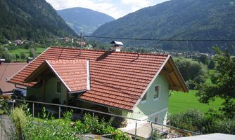 A pretty house with a red tiled roof in a green landscape. In the background, there are mountains and a blue sky.