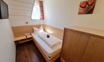 A simple room with a single bed and light wooden furniture. A picture with hearts hangs on the wall, and a window lets in daylight.