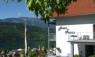 A picturesque house by a lake with green mountains in the background. The sky is clear and sunny, perfect for a relaxing stay.